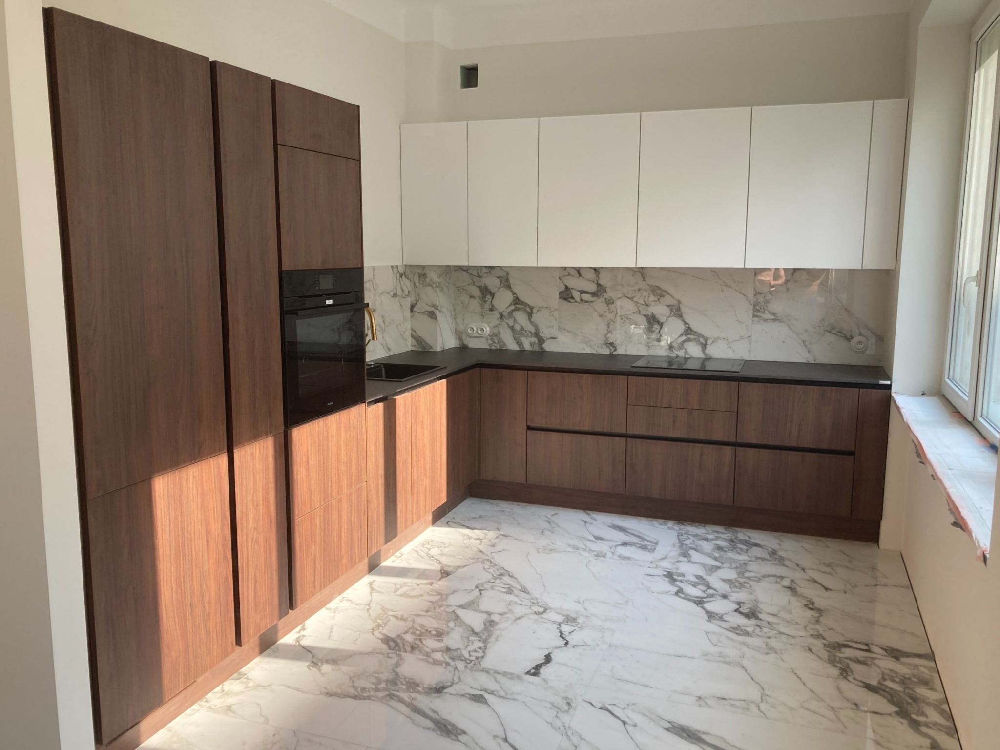 Nowoczesna kuchnia z białymi górnymi szafkami i dolnymi szafkami z ciemnego drewna, czarny blat, marmurowe płytki na ścianie i podłodze, widoczne okno w prawym górnym rogu.