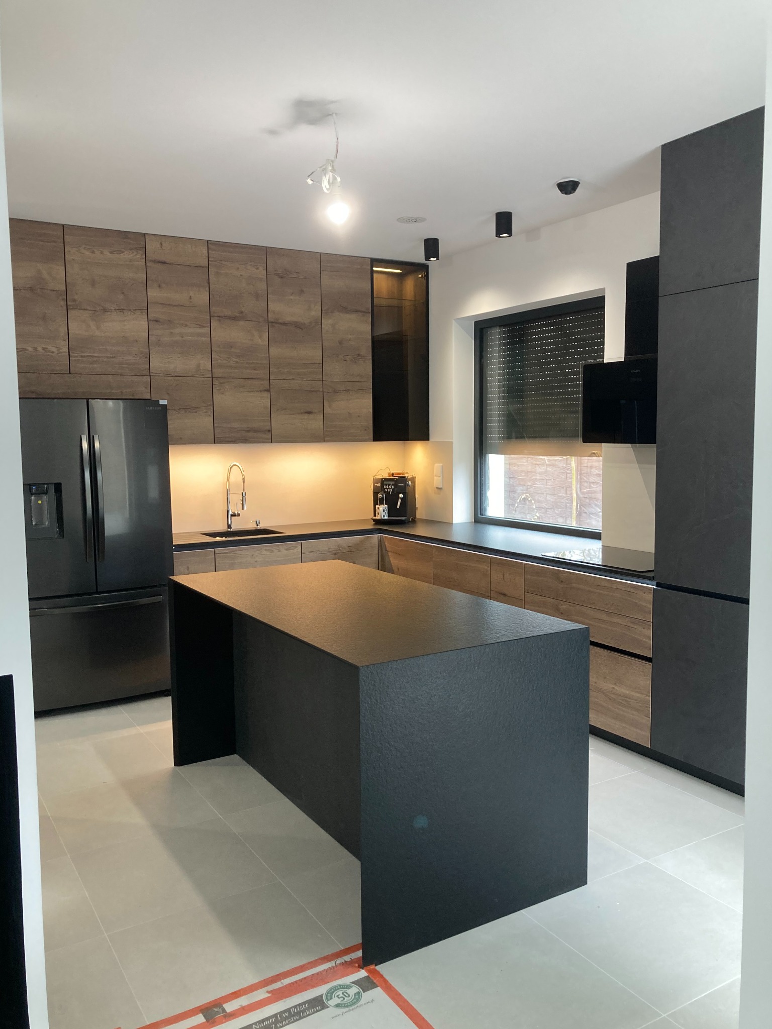Nowoczesna kuchnia z wyspą: połączenie drewnianych frontów szafek górnych i dolnych z czarnym blatem i wyspą, oświetlenie podszafkowe, czarna lodówka side-by-side, ekspres do kawy na blacie.