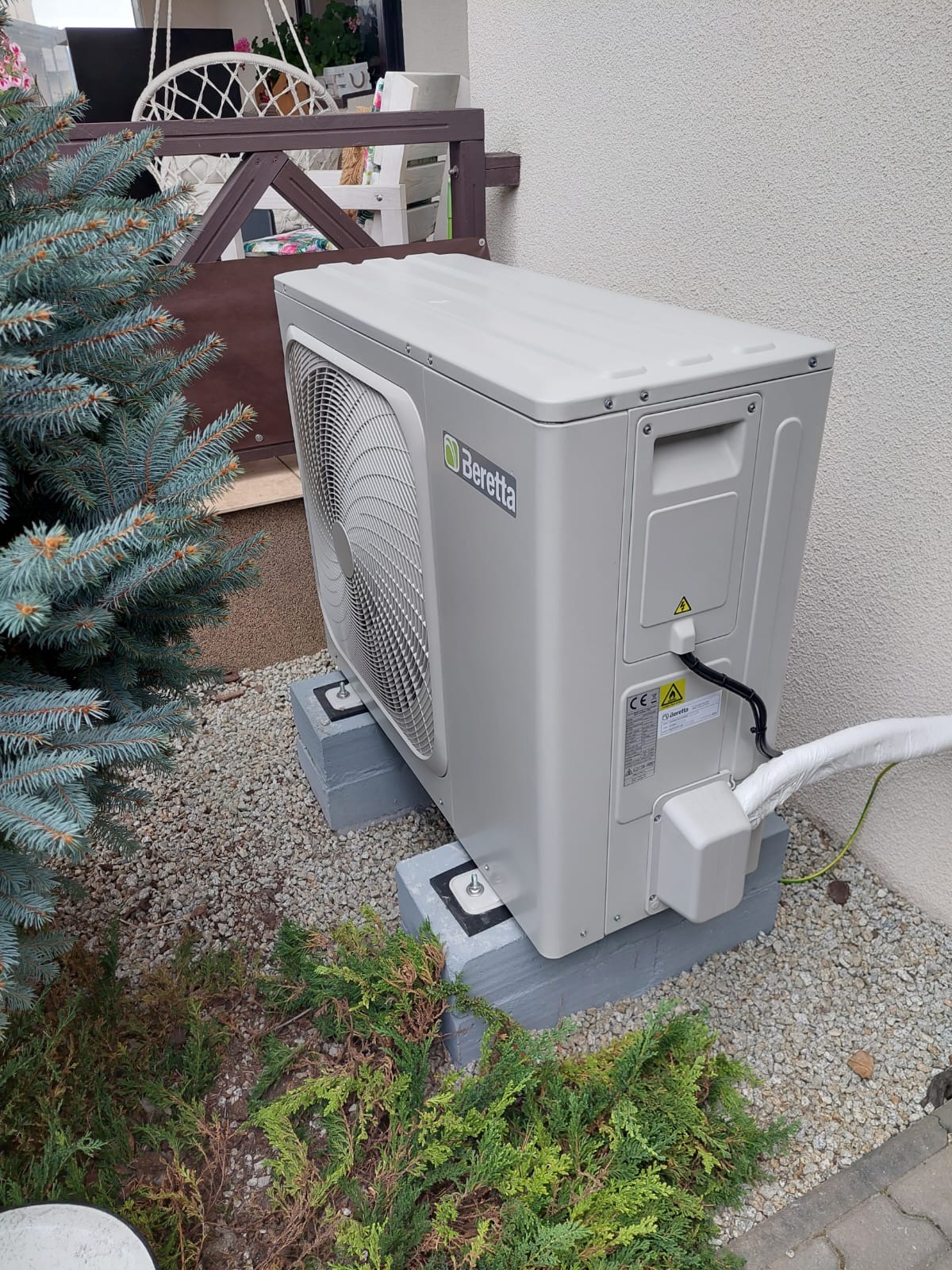Zewnętrzna jednostka klimatyzacji Beretta umieszczona na betonowych podstawach w ogrodzie z szarym żwirem i iglakami.