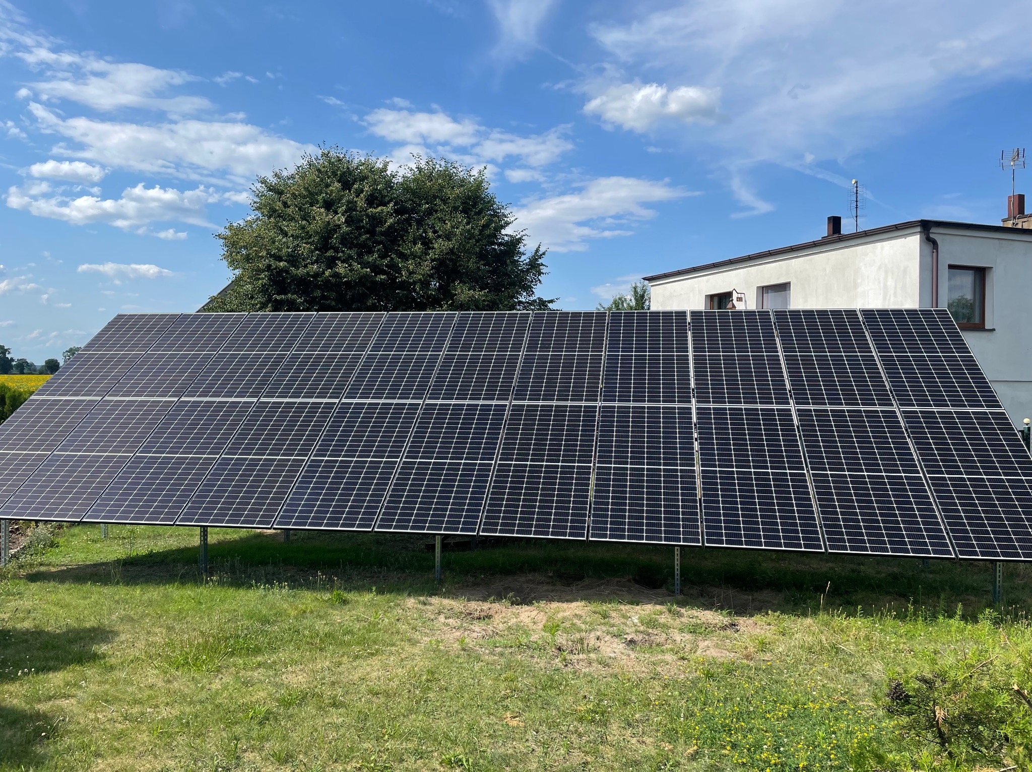 Instalacja paneli słonecznych na metalowej konstrukcji, widok z niskiej perspektywy na tle budynku mieszkalnego i drzewa, błękitne niebo z chmurami w tle.