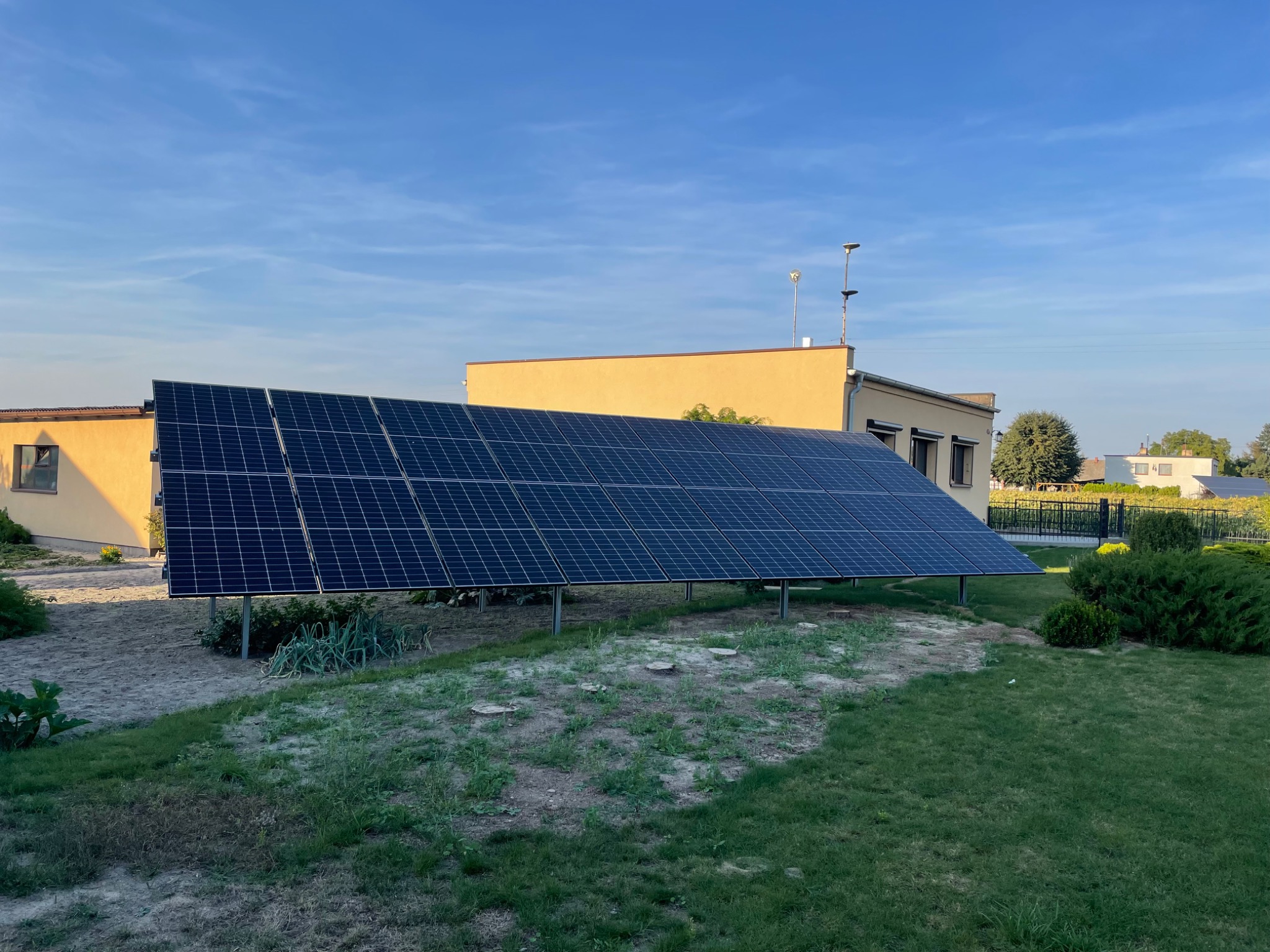 Panel słoneczny zamontowany na metalowych nogach na trawniku przed budynkiem w słoneczny dzień.