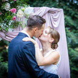 Phost Events - Pocałunek nowożeńców na tle dekoracji ślubnej z różowych tkanin i kwiatów w odcieniach różu i bieli.