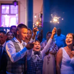 Uśmiechnięci goście weselni trzymają zapalone zimne ognie w sali z niebieskim oświetleniem podczas uroczystości.