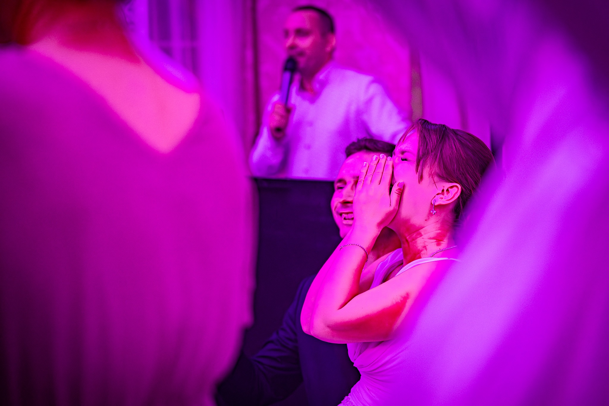 Para młoda reaguje wzruszeniem podczas przemówienia na weselu, oświetlona fioletowym światłem.