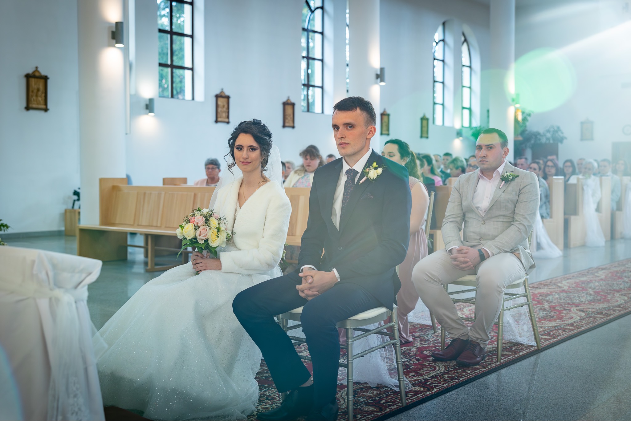 Para młoda siedzi w kościele podczas ceremonii ślubnej, panna młoda trzyma bukiet kwiatów, a za nimi siedzą goście.