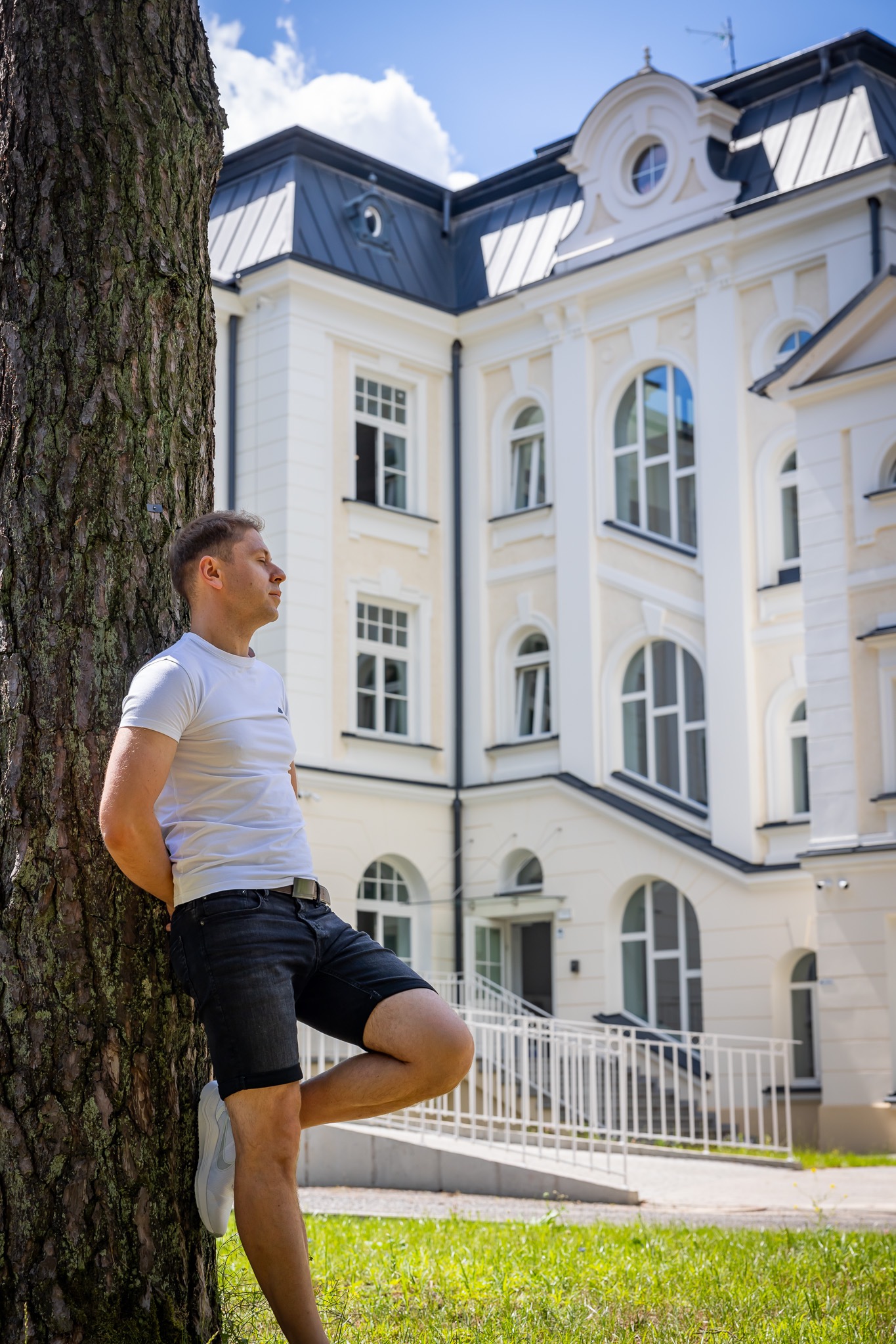 Mężczyzna oparty o drzewo na tle odnowionego, trzypiętrowego budynku w stylu klasycystycznym z jasną fasadą, ciemnym dachem i licznymi oknami z łukami.