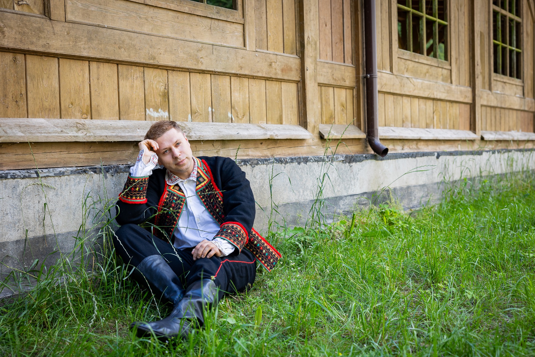 Mężczyzna w stroju ludowym siedzi na trawie oparty o drewniany budynek z oknami.