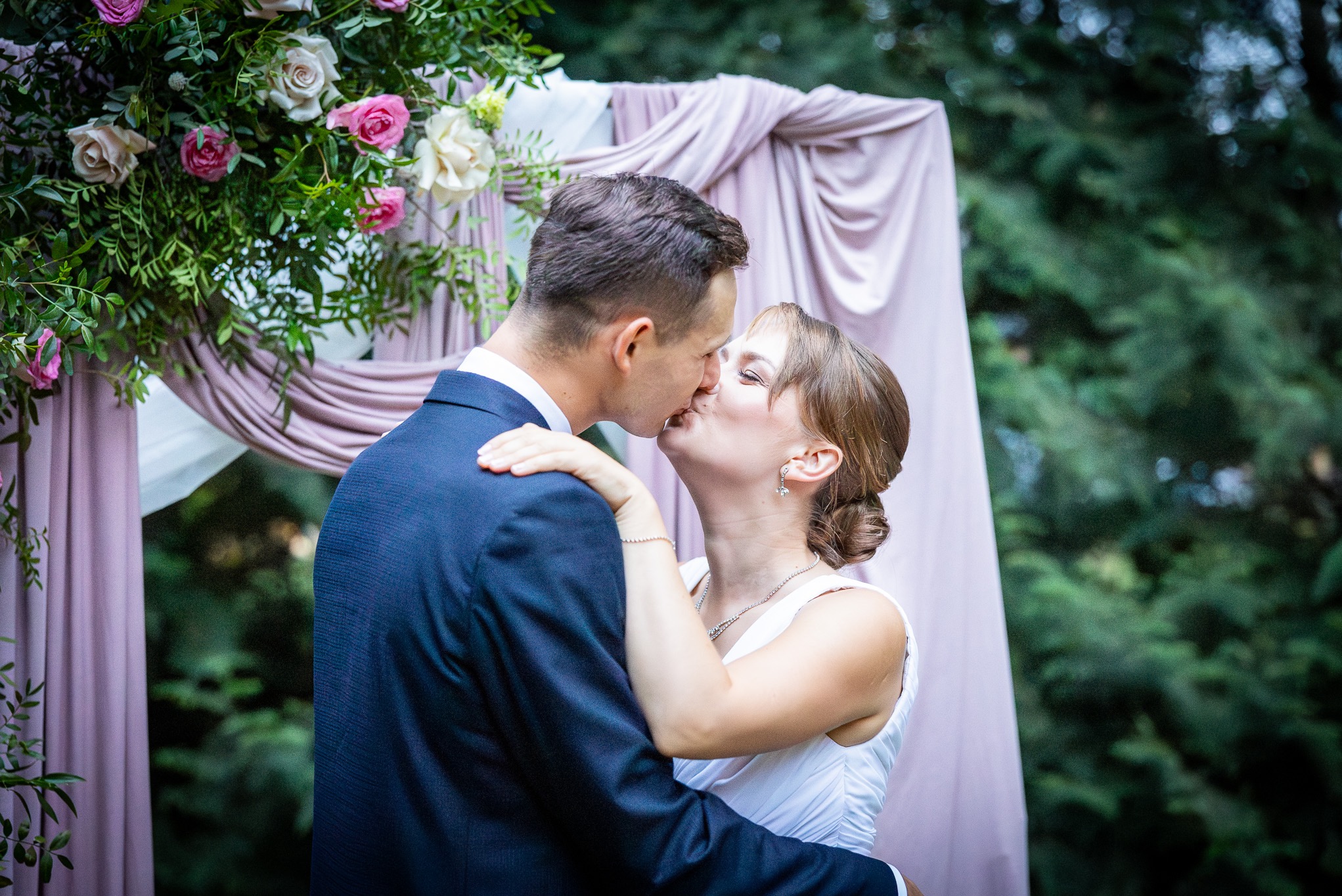 Pocałunek nowożeńców na tle dekoracji ślubnej z różowych tkanin i kwiatów w odcieniach różu i bieli.