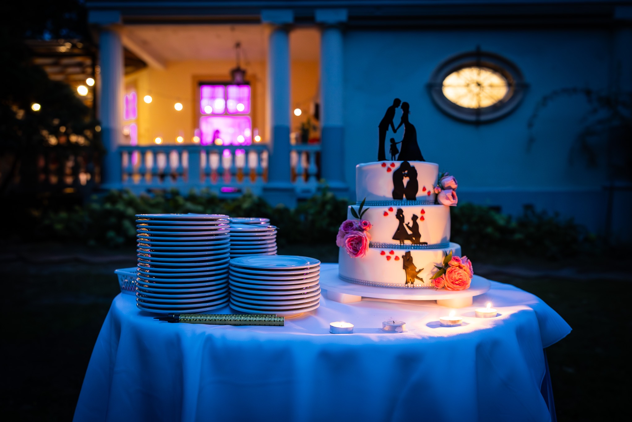 Trzypiętrowy tort weselny z sylwetkami pary młodej i rodziny, zdobiony różami i serduszkami, stoi na stole przykrytym białym obrusem, obok ułożone w stosy talerze, oświetlony świecami, z budynkiem...