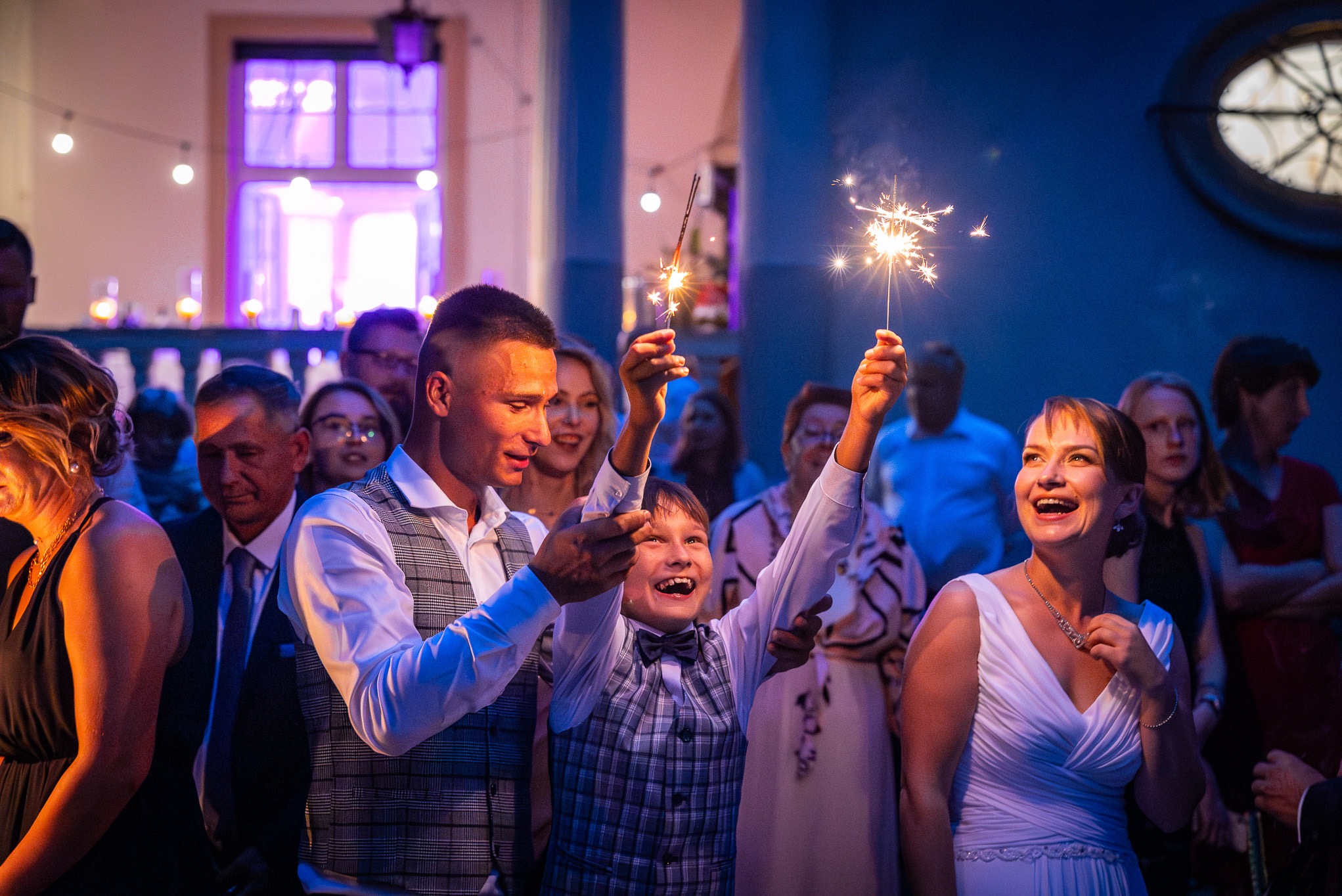 Uśmiechnięci goście weselni trzymają zapalone zimne ognie w sali z niebieskim oświetleniem podczas uroczystości.