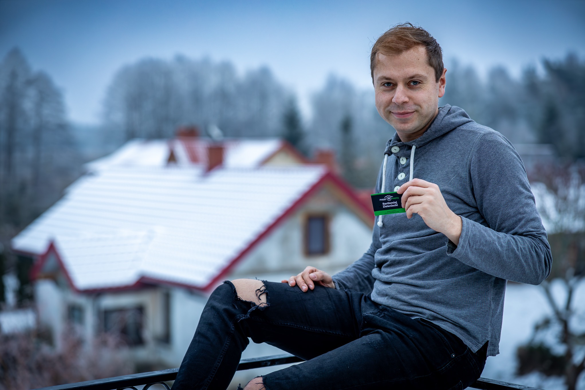 Mężczyzna siedzi na barierce z widokiem na zasypany śniegiem dom, trzymając wizytówkę firmy fotograficznej.