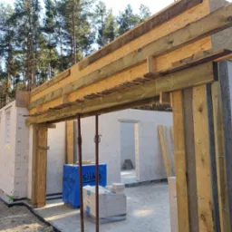 Budowa domu z bloczków silikatowych: widok na wznoszone ściany z otworami okiennymi, drewniane szalunki nad otworem drzwiowym, podpory stalowe, zapakowane bloczki i materiały budowlane, w tle las