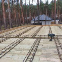 Zbrojenie płyty fundamentowej na deskowaniu z widocznym narzędziem do wiązania drutu zbrojeniowego Makita, w tle las i budynek w stanie surowym otwartym.