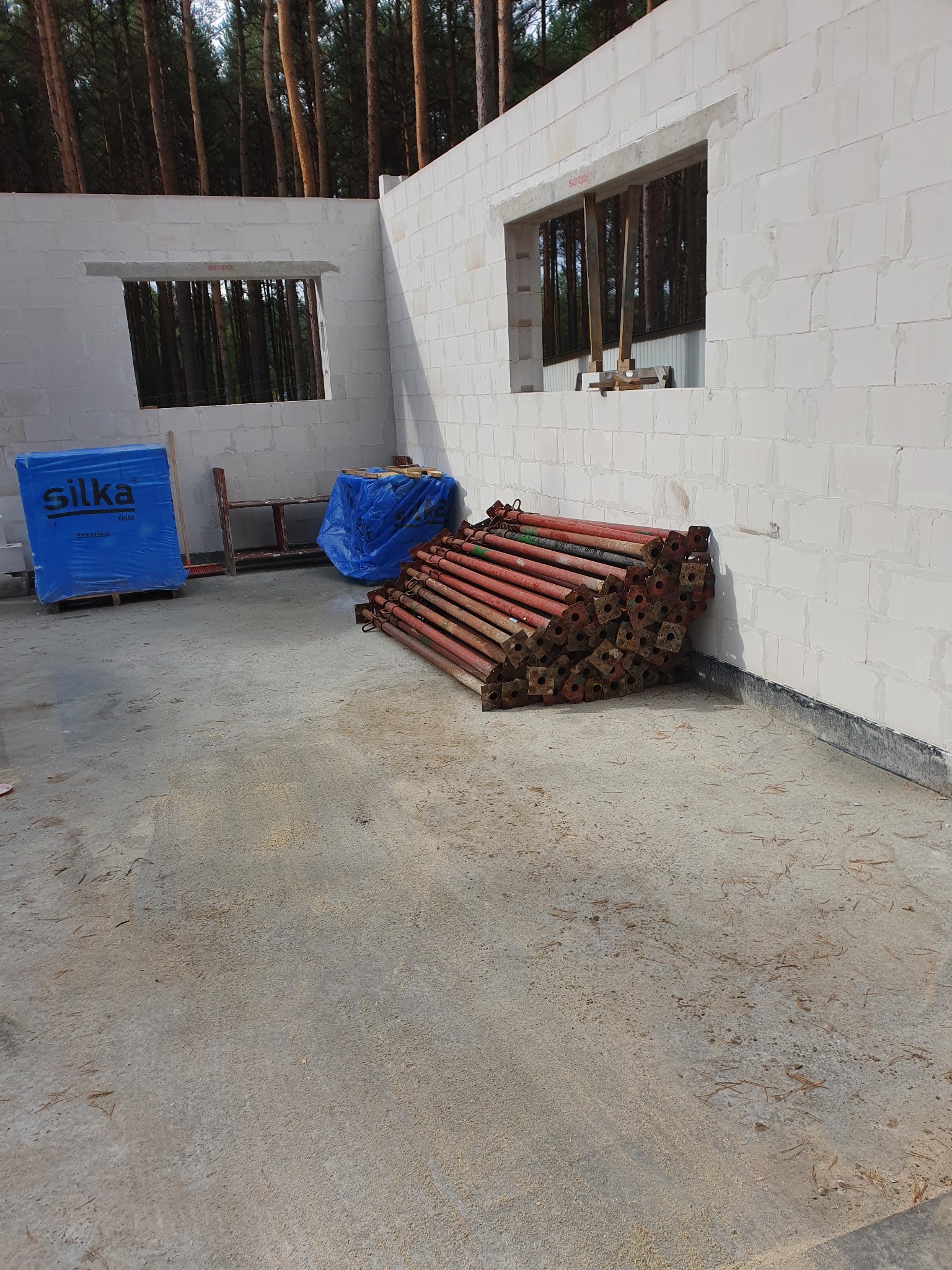 Wnętrze surowego domu w trakcie budowy: ściany z białych bloczków z otworami okiennymi, sterta czerwonych podpór budowlanych, paleta zafoliowanych bloczków Silka, widok na las przez okna.