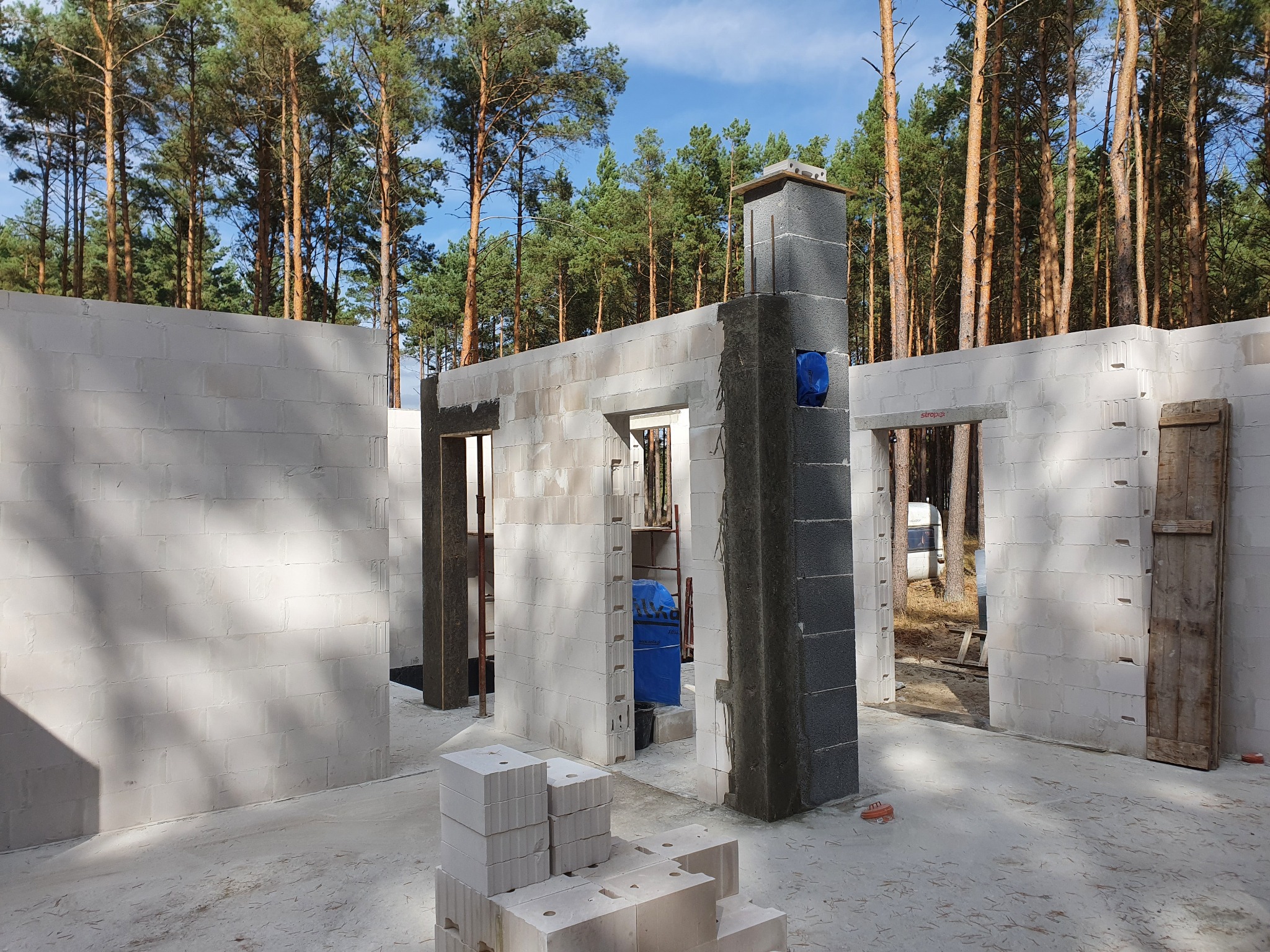 Wnętrze domu w trakcie budowy z białych bloczków, widoczny komin w szarym tynku, w tle las sosnowy i błękitne niebo.