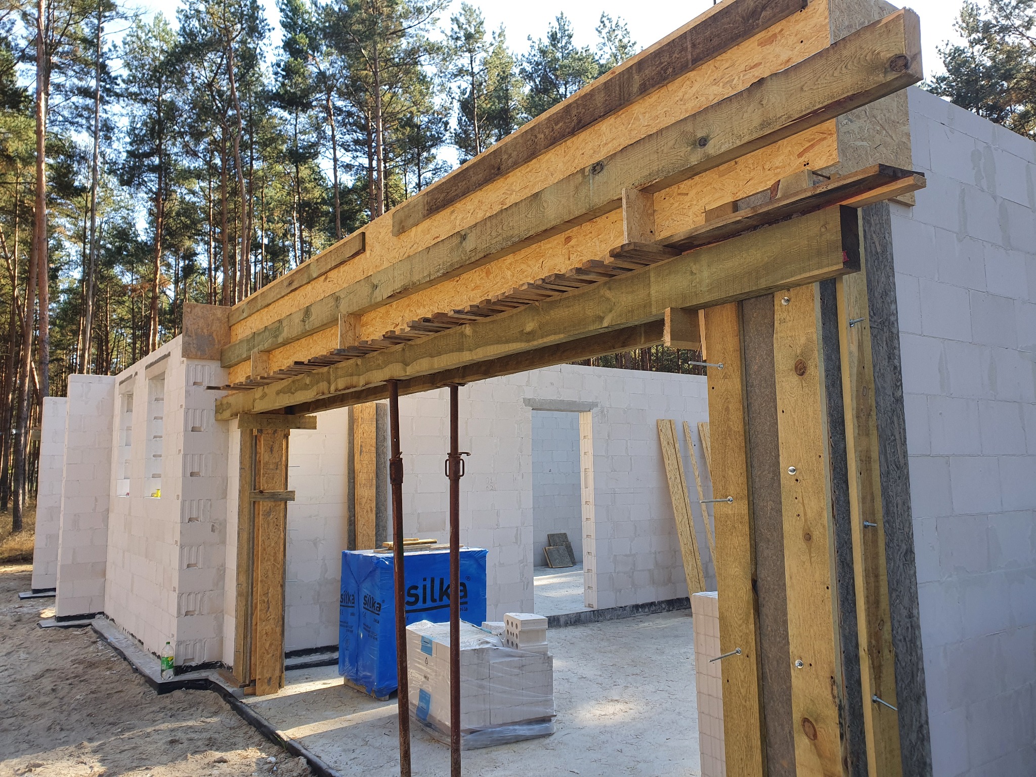 Budowa domu z bloczków silikatowych: widok na wznoszone ściany z otworami okiennymi, drewniane szalunki nad otworem drzwiowym, podpory stalowe, zapakowane bloczki i materiały budowlane, w tle las