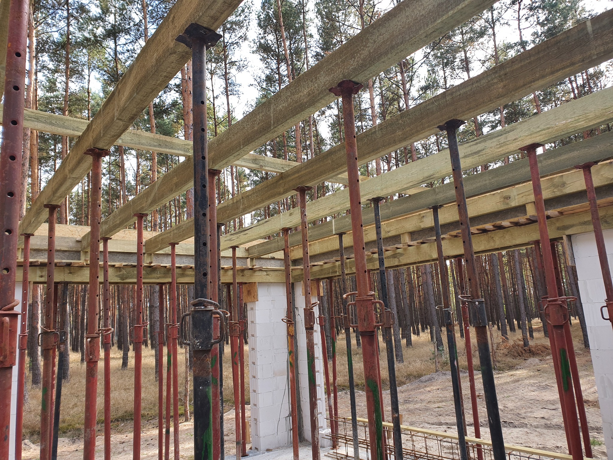 Konstrukcja szalunkowa stropu podtrzymywana przez stemple budowlane na tle lasu sosnowego, widoczne fragmenty muru z bloczków.