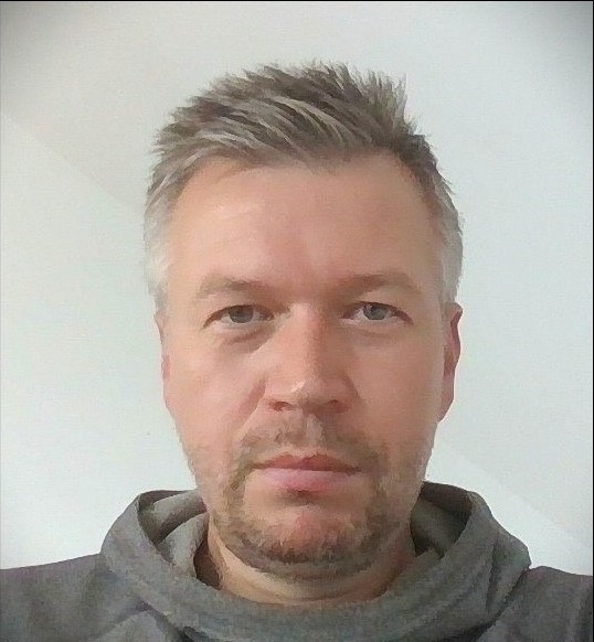 Portret mężczyzny z siwiejącymi włosami i lekkim zarostem, ubrany w szarą bluzę z kapturem, na jasnym tle. Ujęcie od przodu, widoczna twarz i część ramion.