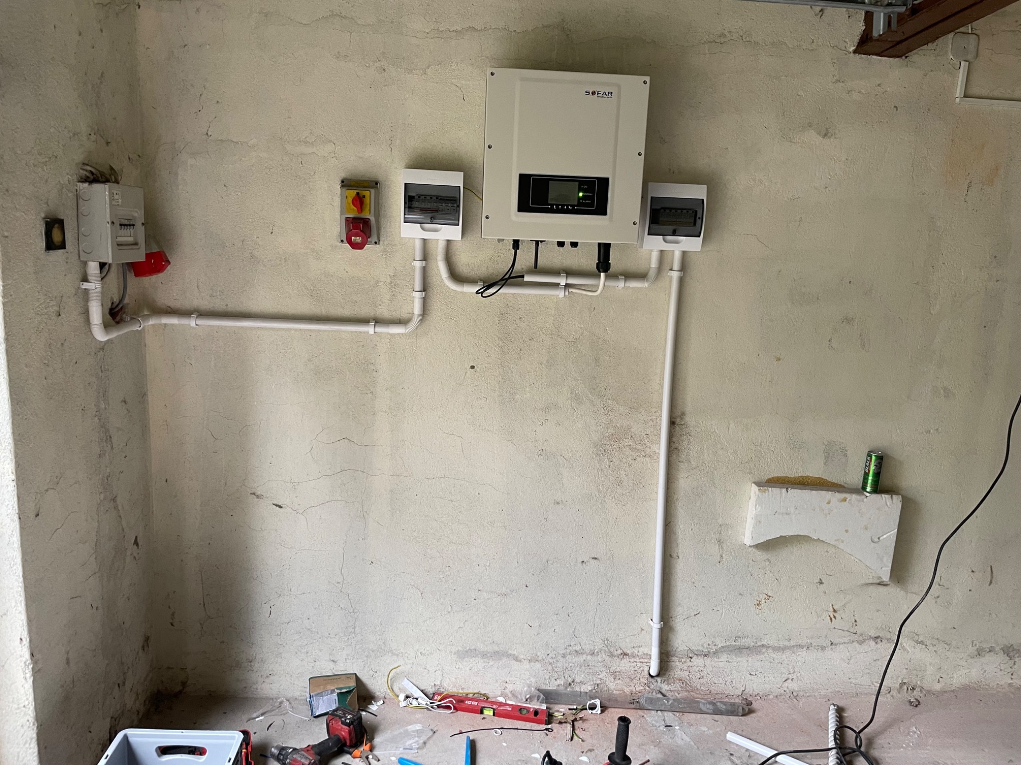 Biały inwerter solarny marki Sofar zamontowany na surowej ścianie, z widocznymi przewodami i skrzynkami elektrycznymi, oraz narzędziami i puszką napoju energetycznego na podłodze.