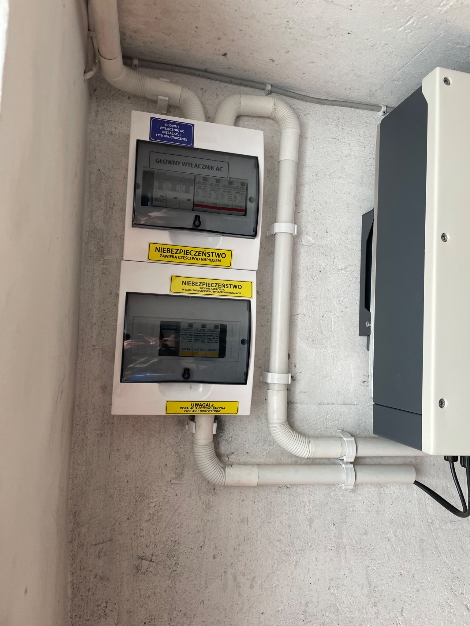 Dwa rozdzielnice elektryczne z naklejkami ostrzegawczymi 'Niebezpieczeństwo' i 'Uwaga! Instalacja fotowoltaiczna' zamontowane na szarej, chropowatej ścianie, z widocznymi białymi rurami...