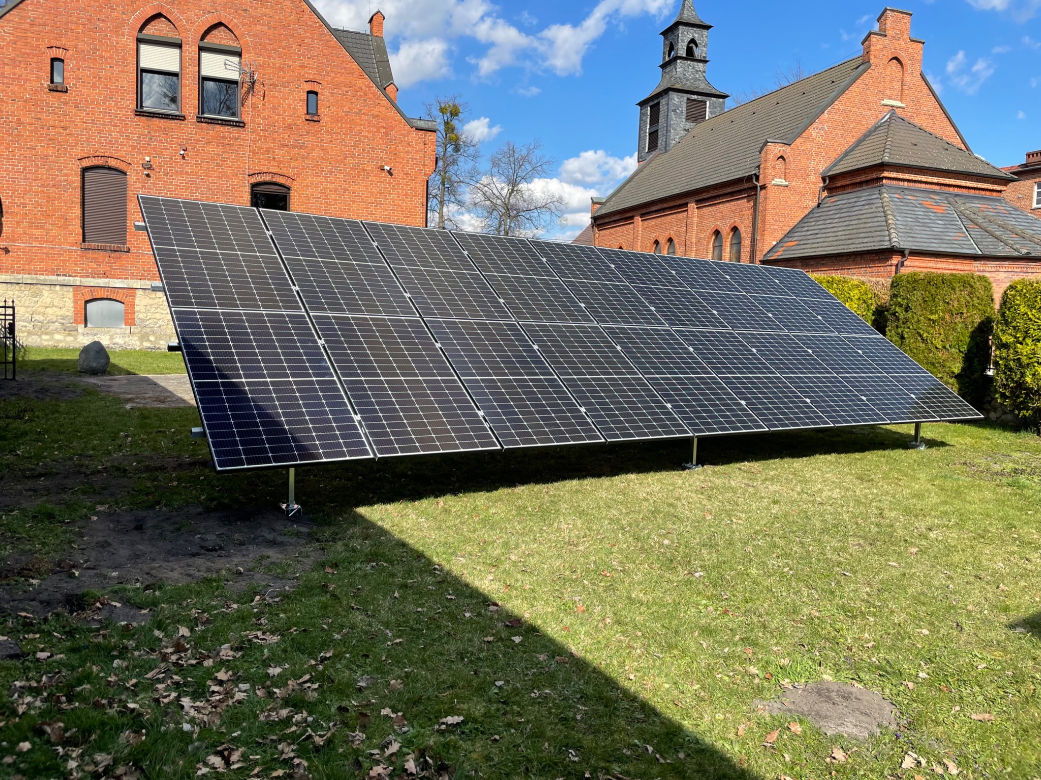 Rząd paneli fotowoltaicznych na trawniku przed ceglanym budynkiem z wieżą kościelną w tle, widoczne mocowania i cień na trawie.
