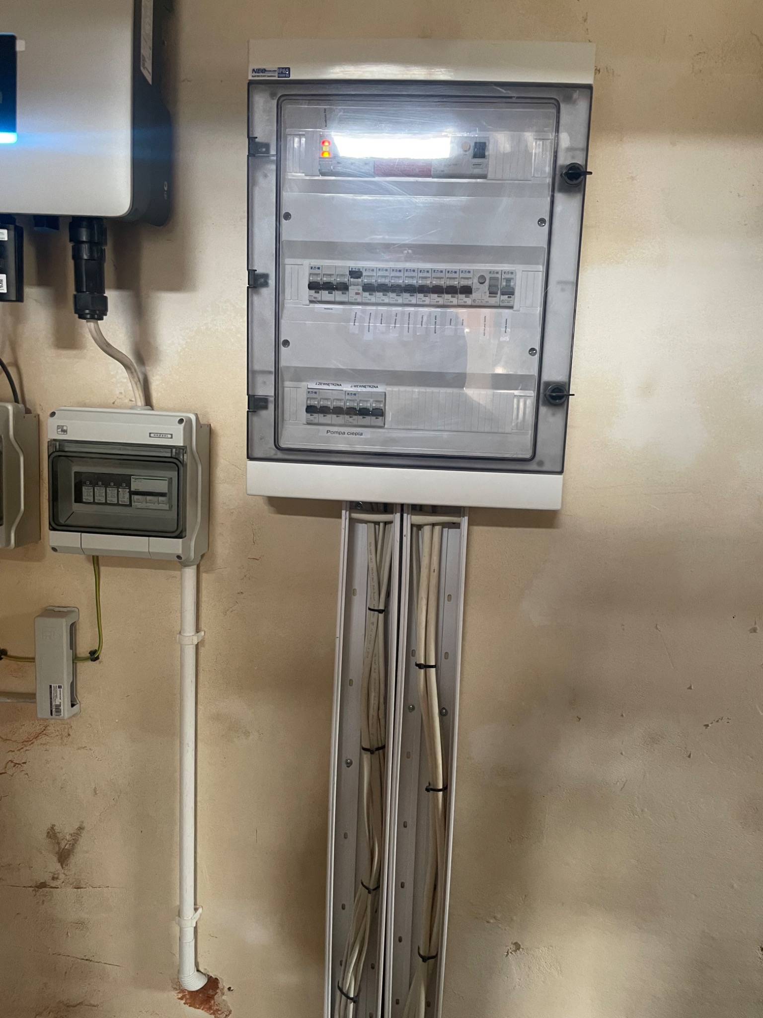 Rozdzielnica elektryczna z przezroczystą obudową, widoczne bezpieczniki i przewody w białych korytkach na beżowej ścianie, obok falownik fotowoltaiczny.
