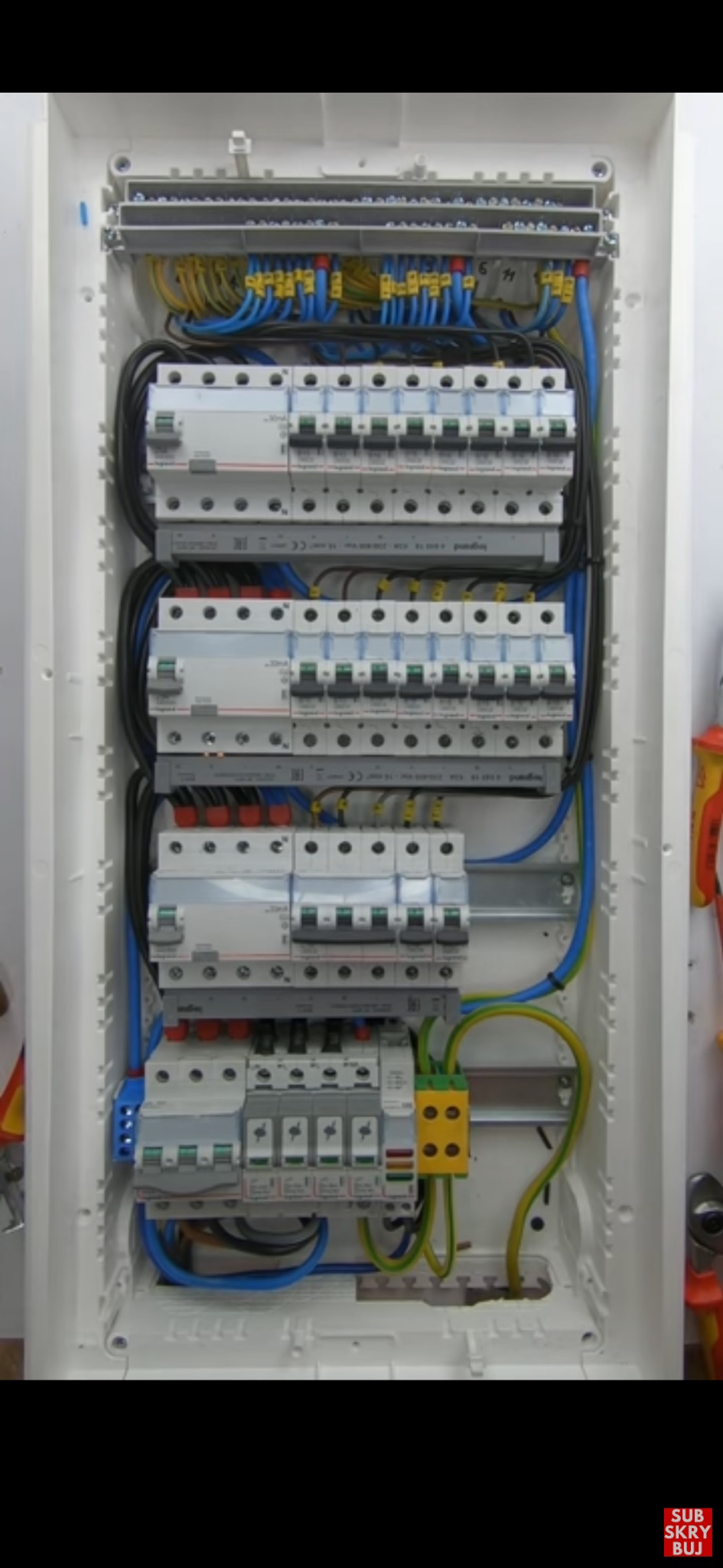 Wnętrze rozdzielnicy elektrycznej z zainstalowanymi bezpiecznikami i okablowaniem w kolorach niebieskim, czarnym, żółto-zielonym i brązowym.