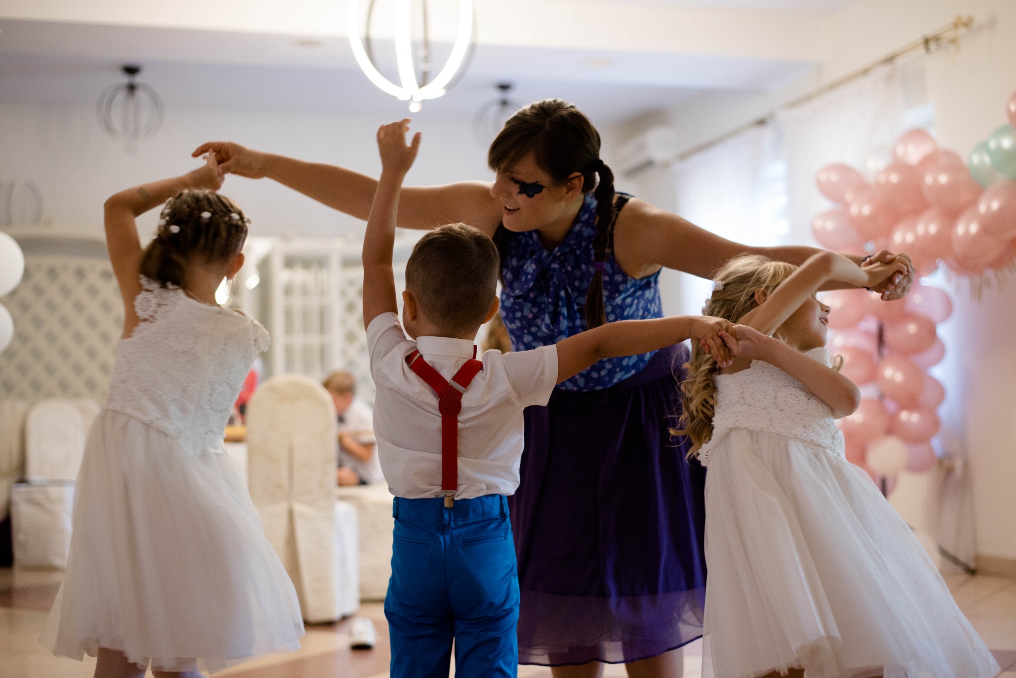 Kobieta z pomalowaną twarzą prowadzi taniec dla dzieci w sukienkach i chłopca w niebieskich spodniach z szelkami na tle balonów i sali bankietowej.
