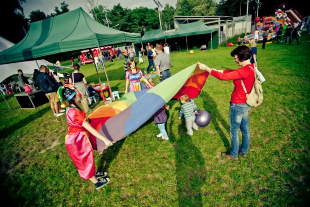 Animator w stroju klauna i kobieta rozciągają kolorową płachtę nad grupą dzieci na trawie podczas imprezy plenerowej, w tle widoczne namioty i dmuchany zamek.