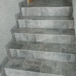 Wykończone schody z szarych płytek ceramicznych o matowej powierzchni, z widocznymi detalami tekstury kamienia i subtelnymi rowkami antypoślizgowymi na krawędziach stopni, przylegające do ściany...