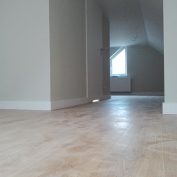 ButMajster - Jasny parkiet w pokoju na poddaszu z białymi listwami przypodłogowymi i ścianami w odcieniach szarości, widoczny fragment okna dachowego i grzejnika.