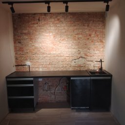 ButMajster - Czarna zabudowa kuchenna z umywalką i baterią, zamontowana na tle ściany z cegły, oświetlona reflektorami sufitowymi, z widocznymi instalacjami hydraulicznymi i elektrycznymi.