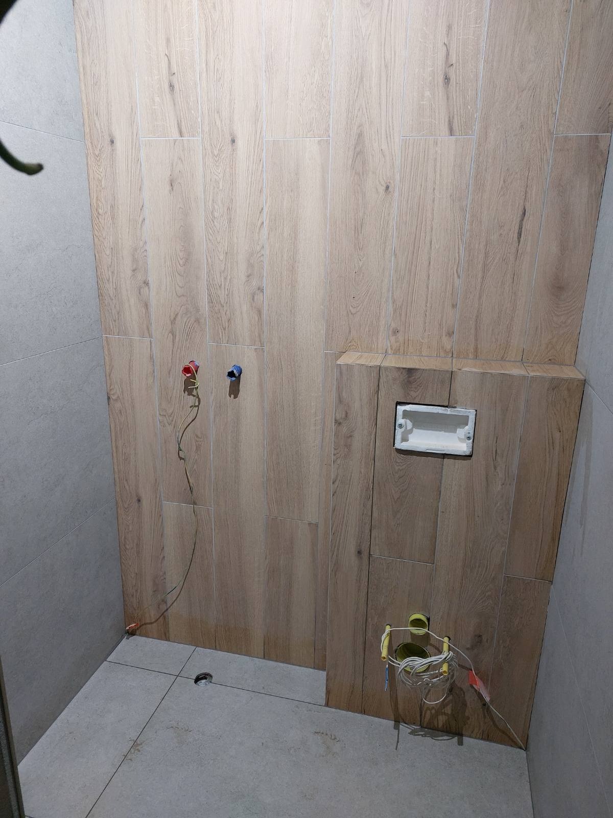 Wnętrze łazienki w trakcie prac: ściany wyłożone płytkami imitującymi drewno, widoczne wystające przewody i rury, szare płytki podłogowe, otwór w podłodze, częściowo zabudowana instalacja...