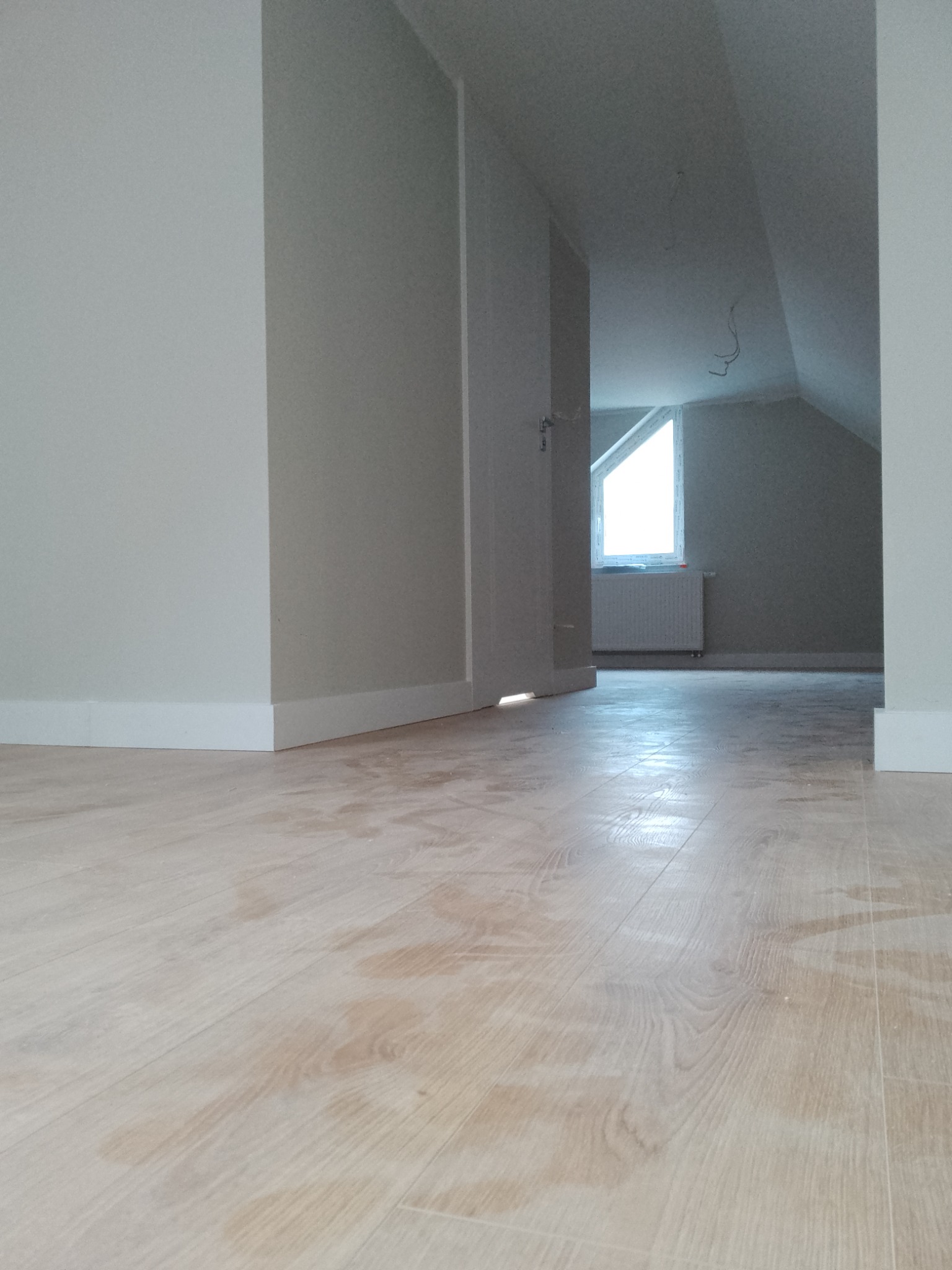 Jasny parkiet w pokoju na poddaszu z białymi listwami przypodłogowymi i ścianami w odcieniach szarości, widoczny fragment okna dachowego i grzejnika.