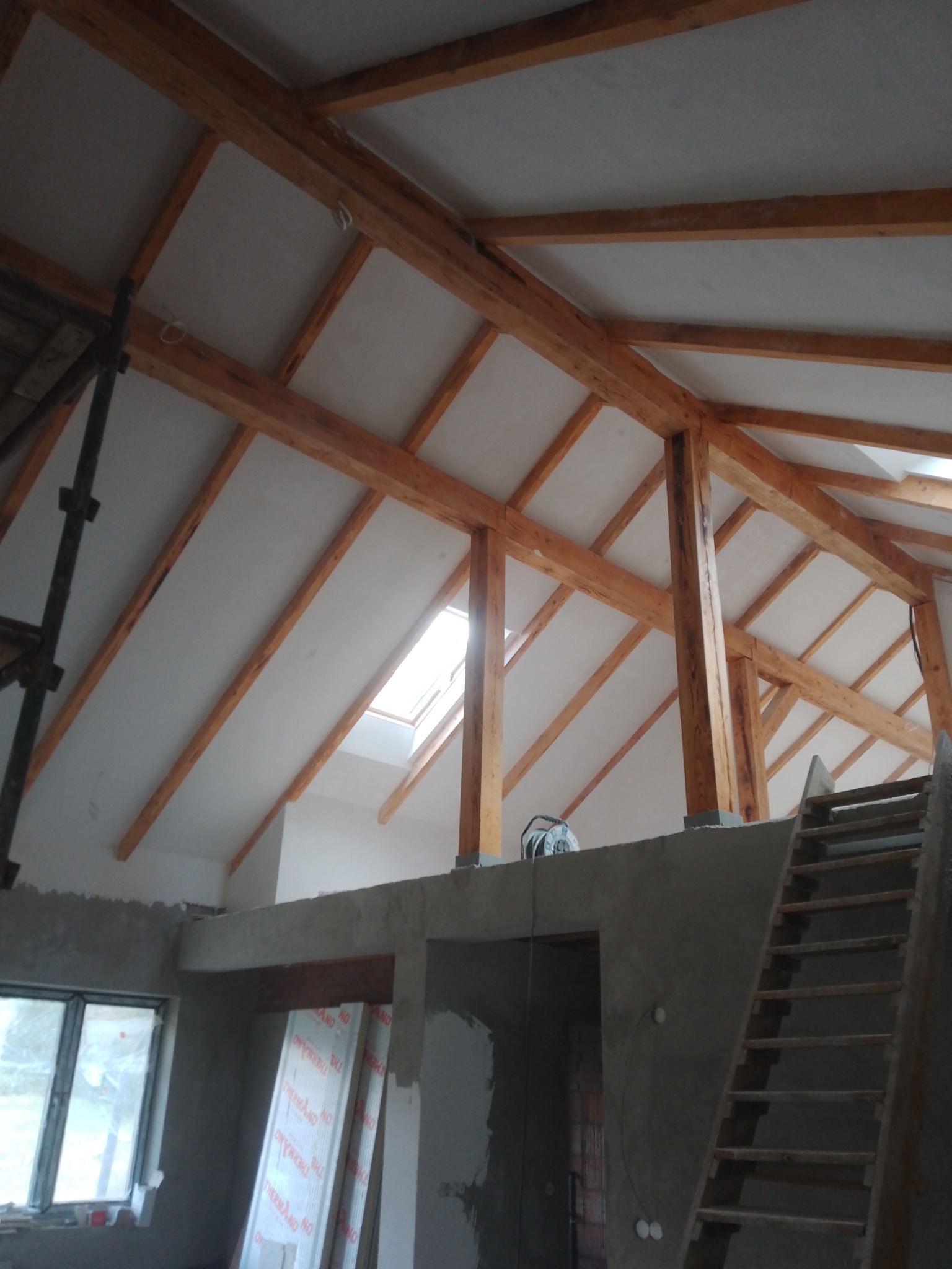 Skośny sufit poddasza z drewnianą konstrukcją nośną i oknem dachowym, częściowo wykończony płytami, widoczne niedokończone ściany z szarą zaprawą i schody drewniane.