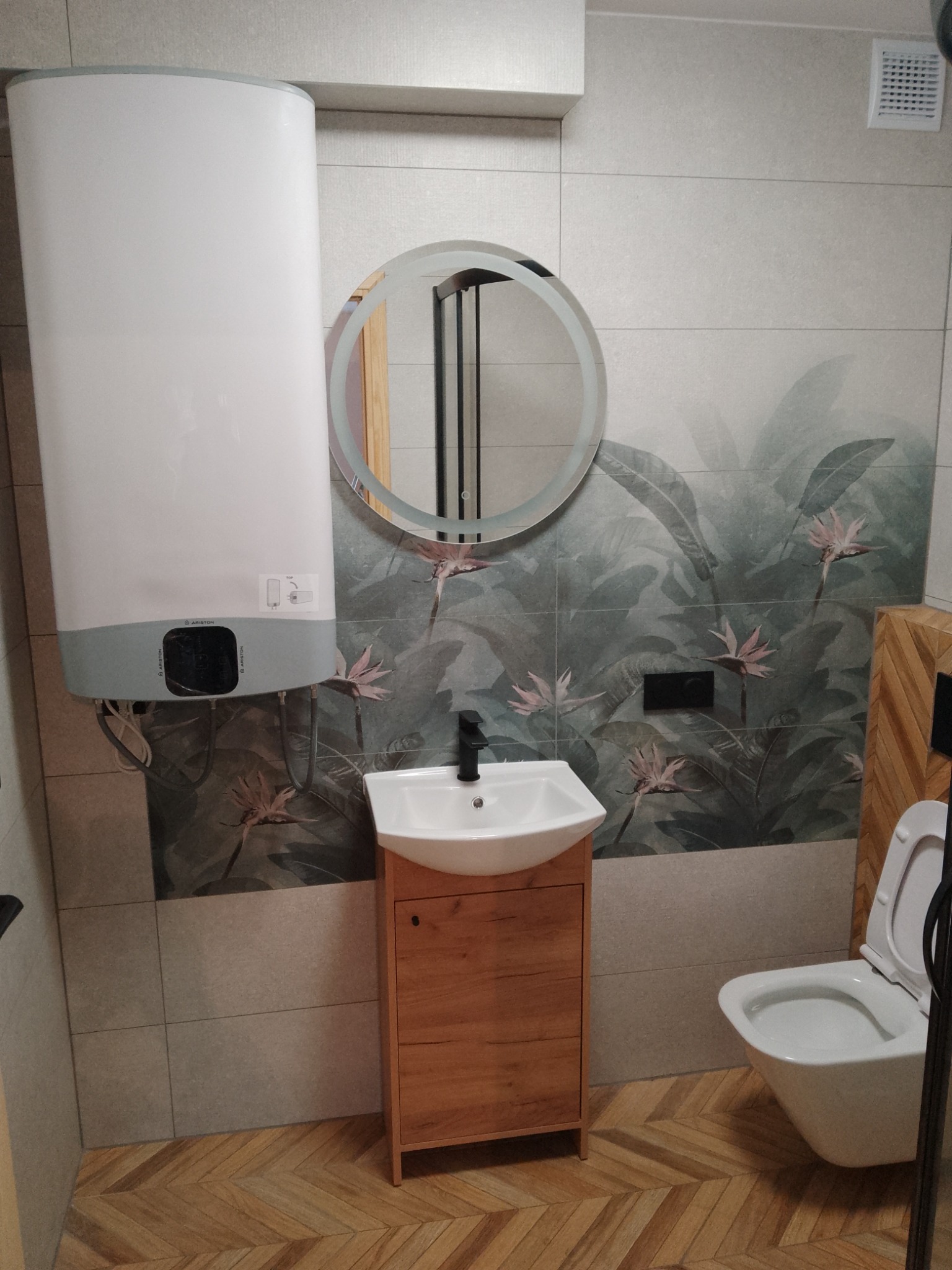 Wykończona łazienka z bojlerem Ariston, okrągłym lustrem z podświetleniem LED, umywalką nablatową na drewnianej szafce i wiszącą toaletą, płytki z motywem tropikalnych kwiatów na ścianach i parkiet...
