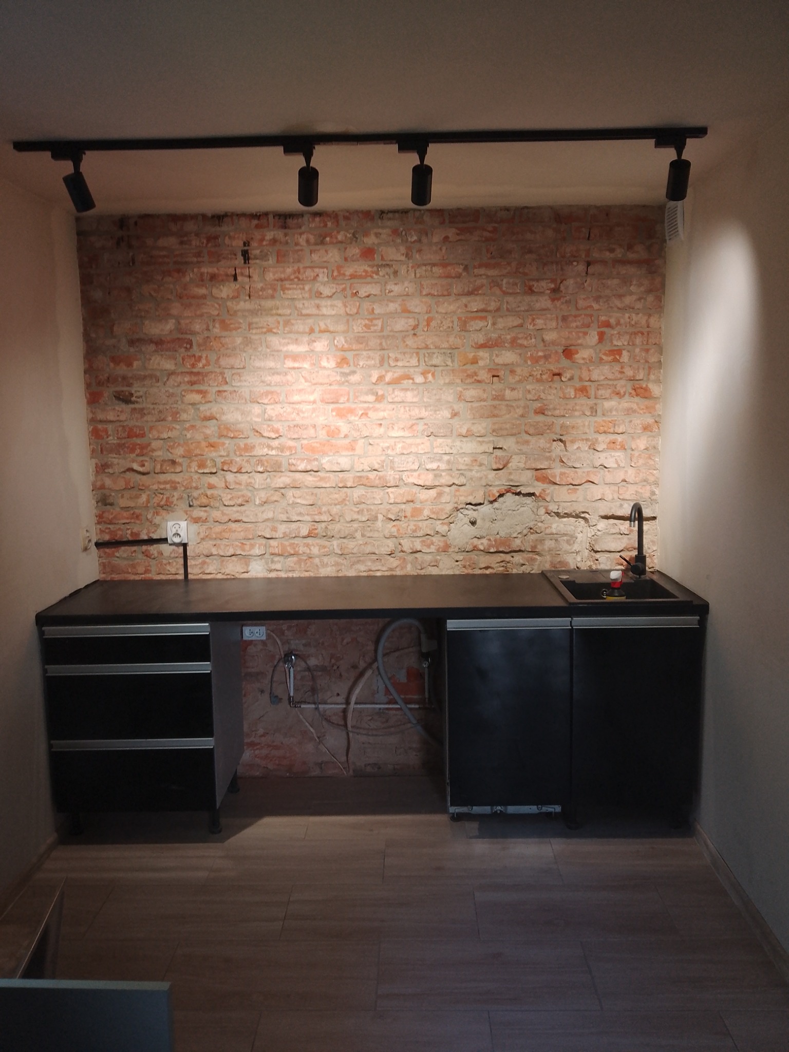 Czarna zabudowa kuchenna z umywalką i baterią, zamontowana na tle ściany z cegły, oświetlona reflektorami sufitowymi, z widocznymi instalacjami hydraulicznymi i elektrycznymi.