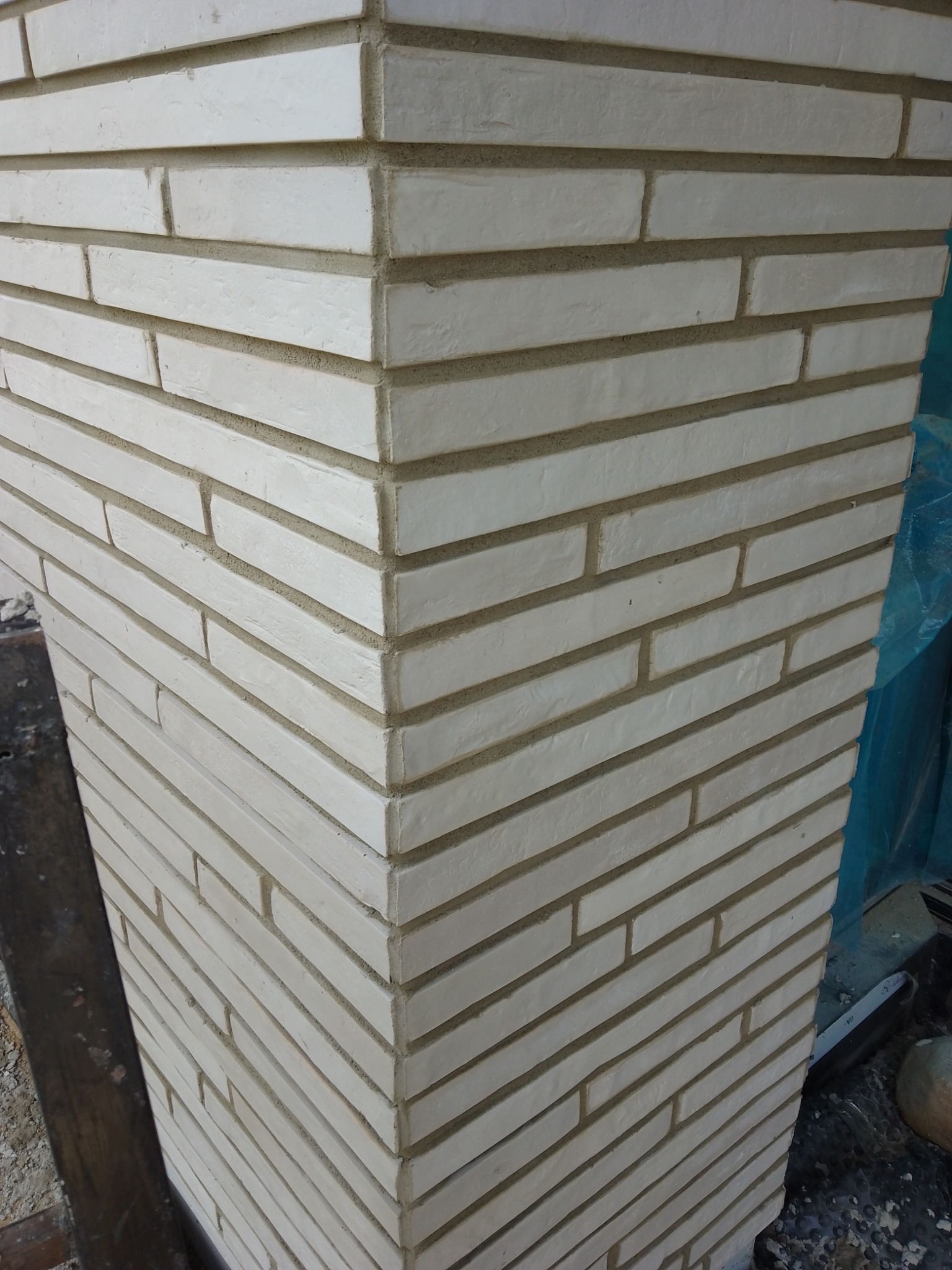 Wykończony narożnik ściany obłożony wąskimi, beżowymi płytkami imitującymi cegłę, z widoczną fakturą i fugami, uchwycony pod kątem.