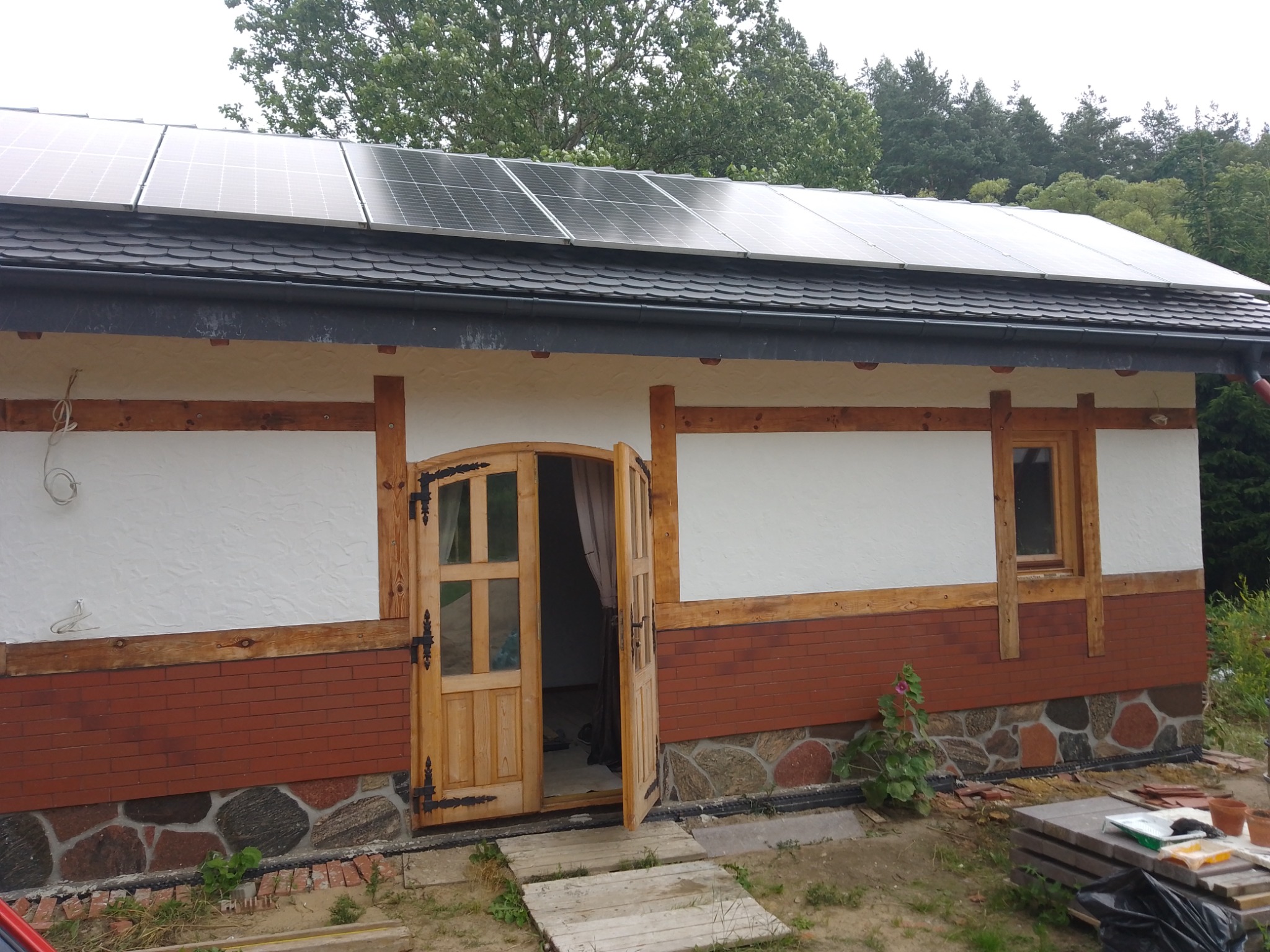 Budynek z panelami słonecznymi na dachu, drewnianą stolarką okienną i drzwiową, oraz elewacją z białego tynku, cegły i kamienia polnego. Drzwi wejściowe są otwarte.