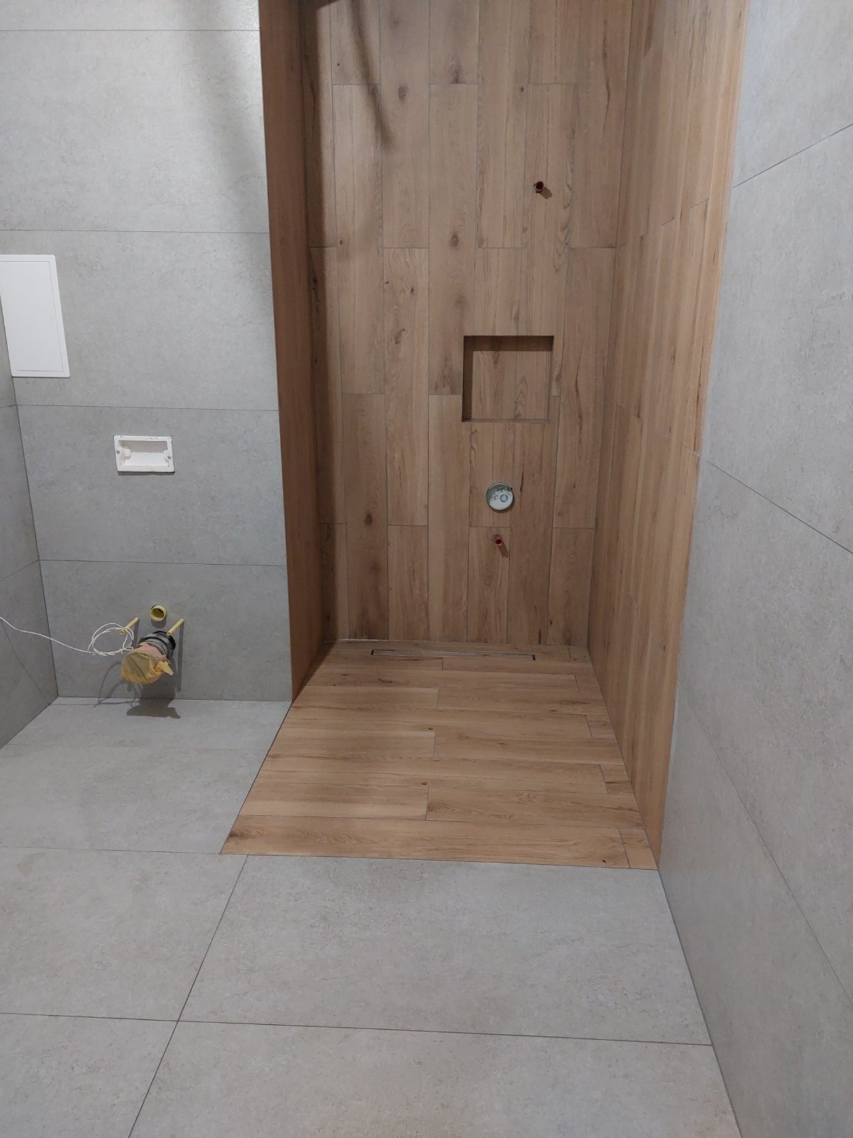 Niedokończona kabina prysznicowa z drewnopodobnymi płytkami na ścianach i podłodze, widoczne przyłącza wodne i odpływ liniowy, szare płytki na pozostałych ścianach i podłodze, narzędzia przy ścianie.