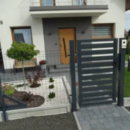 Nowoczesna posesja z grafitową bramą wjazdową z poziomymi panelami, skrzynką na listy i siatką ogrodzeniową, widok z kostki brukowej na tle domu z numerem 8.