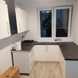 logunoff.pl - Nowoczesna kuchnia w kształcie litery U z białymi szafkami, ciemnym blatem i drewnopodobną podłogą; widoczny karton na podłodze sugerujący niedawny montaż.