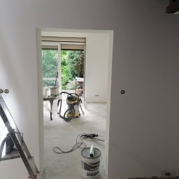 logunoff.pl - Wnętrze w trakcie odświeżania: ściany pomalowane na szaro, widoczny otwarty pojemnik z farbą Profi, odkurzacz przemysłowy, drabina aluminiowa i narzędzia malarskie na podłodze. Okno z widokiem...