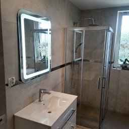 logunoff.pl - Nowoczesna łazienka z kabiną prysznicową, podświetlanym lustrem i białą szafką z umywalką, wykończona szarymi płytkami imitującymi kamień.