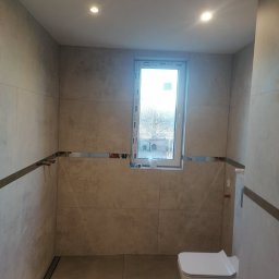 logunoff.pl - Wykończona łazienka z szarymi płytkami imitującymi kamień, minimalistyczna toaleta podwieszana, okno z widokiem na drzewa, chromowane listwy dekoracyjne i odpływ liniowy w podłodze.