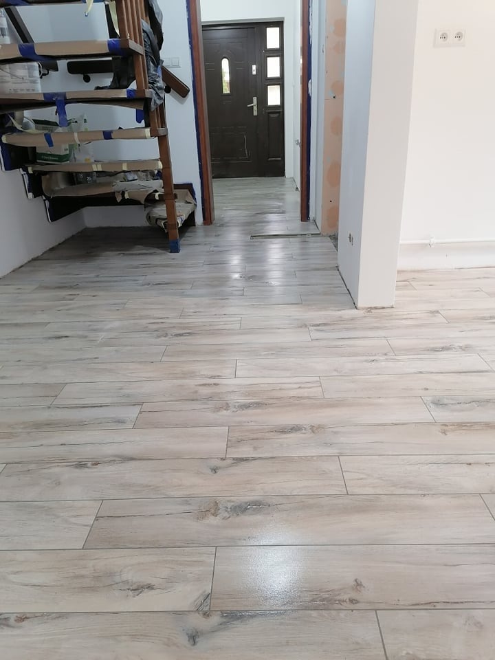 Ułożona podłoga z płytek imitujących drewno w odcieniach beżu i szarości, widoczny fragment zabezpieczonych schodów drewnianych i drzwi wejściowych.