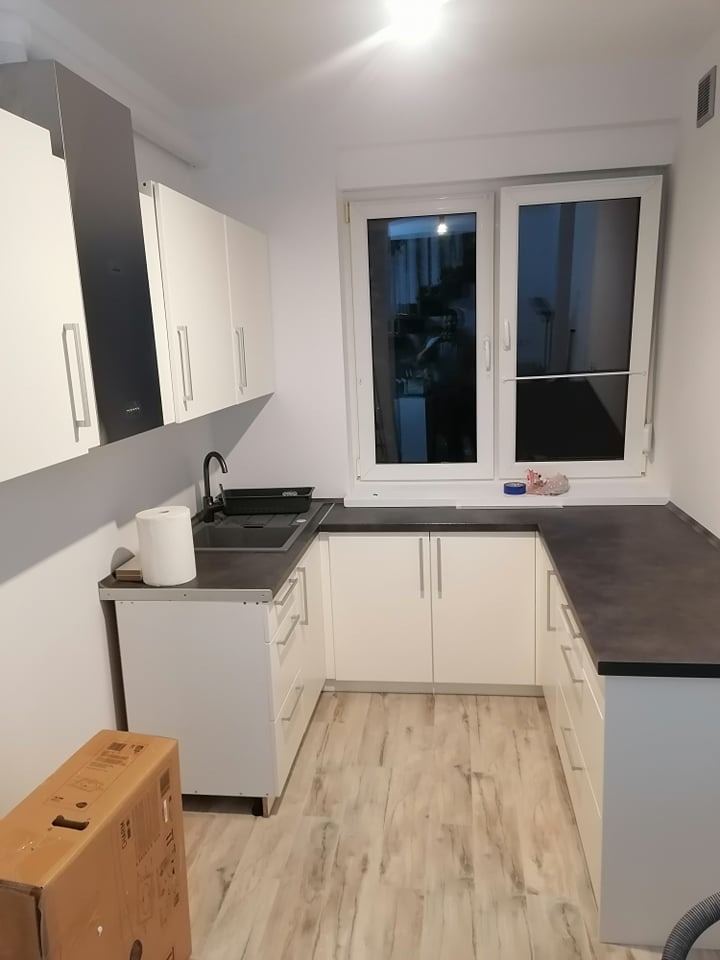 Nowoczesna kuchnia w kształcie litery U z białymi szafkami, ciemnym blatem i drewnopodobną podłogą; widoczny karton na podłodze sugerujący niedawny montaż.