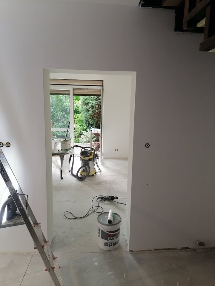 Wnętrze w trakcie odświeżania: ściany pomalowane na szaro, widoczny otwarty pojemnik z farbą Profi, odkurzacz przemysłowy, drabina aluminiowa i narzędzia malarskie na podłodze. Okno z widokiem...