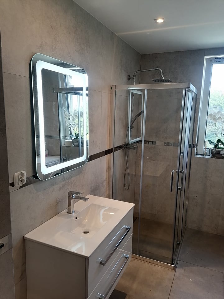 Nowoczesna łazienka z kabiną prysznicową, podświetlanym lustrem i białą szafką z umywalką, wykończona szarymi płytkami imitującymi kamień.