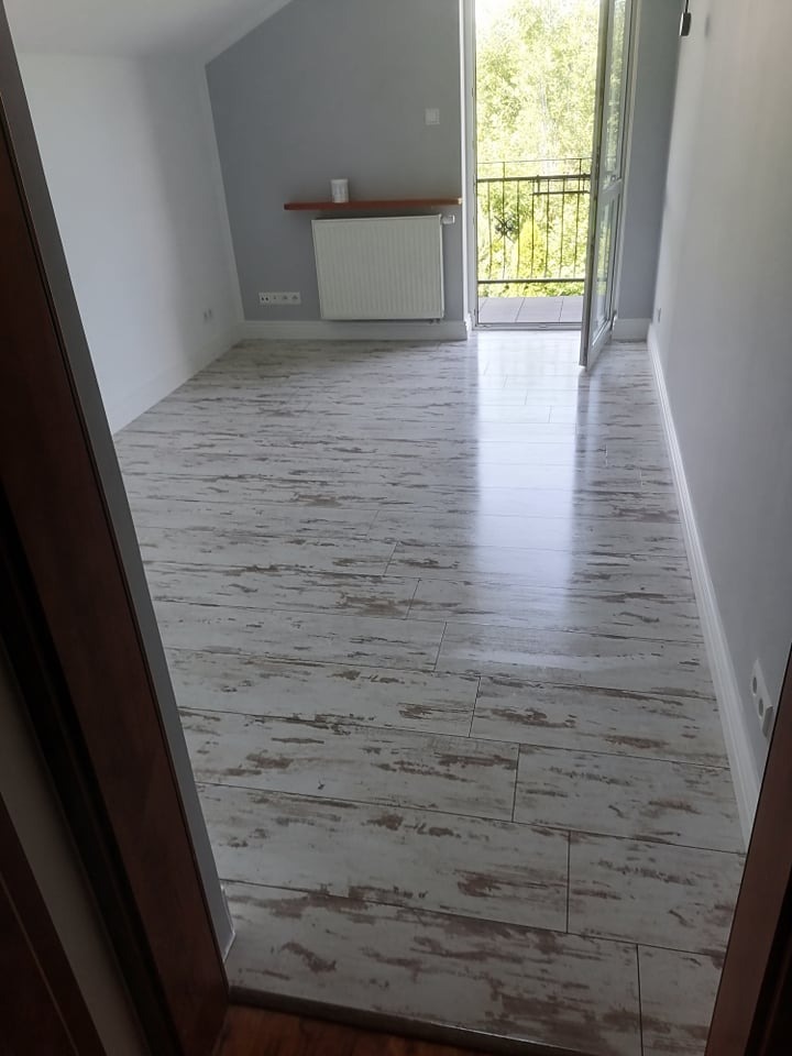 Jasne wnętrze poddasza z podłogą imitującą postarzane drewno, widok na balkon przez otwarte drzwi balkonowe.